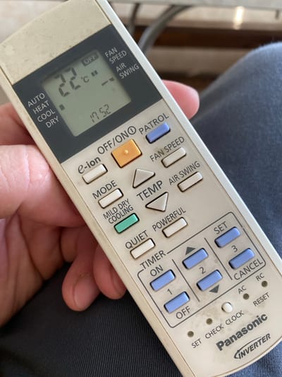 A close-up shot shows a person's hand holding a beige and grey Panasonic air conditioner remote control. The remote has various buttons, including "ON," "OFF," "TIMER," "MODE," "FAN SPEED," "AIR SWING," and directional arrows. The display screen shows "22 C" and a time of "17:52." The remote is positioned against a background of dark blue fabric, possibly clothing. The lighting suggests it is daytime, but there are no clear indicators of weather or a specific location beyond the fabric. The subjects are the hand and the remote control. The activity is holding and likely operating the remote. Notable details include the visible text on the remote, the temperature reading, and the time. The context provided about the location in Belarus is not visually evident in the image itself.