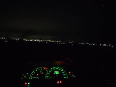 The image displays a nighttime perspective from within a stationary vehicle overlooking the city of Balsha, Bulgaria. The lower third of the frame shows the illuminated dashboard, featuring a tachometer registering approximately 0-1000 RPM and a speedometer at 0 km/h. A digital display within the speedometer reads "28" and "135832". The fuel gauge indicates a low level, and the engine temperature gauge shows a cold engine. Active warning indicators include an orange engine symbol and a red battery symbol. Additional red and green indicator lights are visible along the bottom edge of the dashboard. Above the dashboard, the distant lights of the city form a horizontal glow against a dark night sky. A dark, bare tree branch is faintly discernible in the midground against the city lights on the left side of the frame. No individuals are present in the scene.