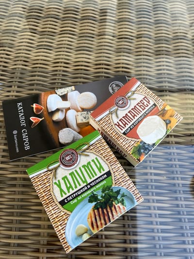 The image shows two boxes of cheese and a catalog placed on a wicker-patterned surface, likely a table. The catalog, on the left, is dark with text in Russian and images of various cheeses. The box on the right, with a red and beige design, features the text "КАМАЛБЕР" and an image of a round white cheese. The box in the foreground, with a green and beige design, displays the text "ХАЛУМИ" and an image of grilled cheese with garlic and parsley. There are no people visible in the image, and the setting does not provide any specific information about the location within Akhlebaikha (Kovriginskii S/S), Russia.