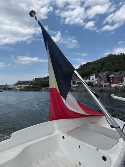 The image shows a white boat moving on a river, with a flag flying from a pole. The flag has three vertical stripes: black, white, and red, from top to bottom. The boat is in the foreground, and the river flows behind it. In the background, there is a city with buildings lining the riverbank, and a hill covered with trees. The city appears to be Dinant, Belgium, known for its riverside location and fortified citadel. The sky is partly cloudy. There are no people visible in the image.