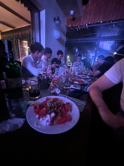 A dimly lit, indoor dining scene in Panagyurski kolonii, Bulgaria, shows a group of people seated at a long table. The table is covered with bottles of wine, water, and Coca-Cola, glasses, and dishes of food, including a plate of what appears to be tomatoes, feta cheese, and herbs. People of varying ages are engaged in conversation. A balloon and security camera are visible on the ceiling. Christmas lights adorn the window.