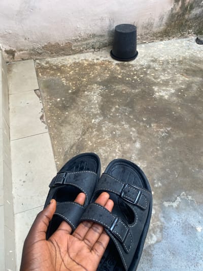 A pair of black sandals is held by a person's hand, with the sandals positioned on a textured, greyish-white tiled surface. The sandals have two straps with buckles, and the stitching is white. To the left of the sandals, on a rougher, stained concrete surface, is a black cylindrical object, possibly a pot or a container. The scene appears to be outdoors, given the natural lighting and the weathered appearance of the surfaces. The overall impression is of a casual, everyday setting. There is no visible text in the image.