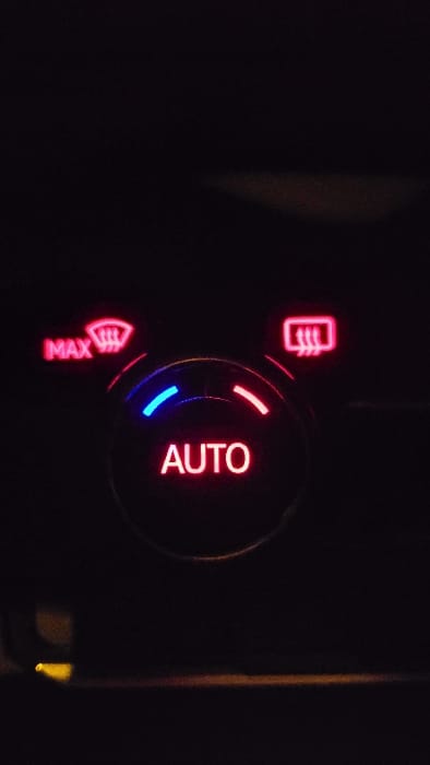 The image displays an illuminated vehicle climate control panel in a dark environment, purportedly located in Magaluf, Spain. A central circular dial features the word "AUTO" in red illumination. An arc of blue light is visible on the upper-left perimeter of this dial, and an arc of red light is on the upper-right. Above and to the left of the central dial, a red-illuminated button or indicator shows a windshield defog symbol with the text "MAX." Above and to the right, another red-illuminated button or indicator displays a rear defog symbol. The overall scene is dark, with only the control panel elements emitting light. A faint yellow light source is visible in the lower-left corner, and a subtle horizontal line of orange light is present along the bottom edge of the frame. No individuals or specific actions are depicted.