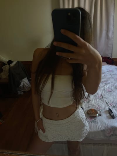 A person is taking a mirror selfie in a bedroom. The person is wearing a white strapless top and a white lace skirt, with a belly button piercing. They have long brown hair and are holding a smartphone to take the photo. In the background, a bed with a floral patterned comforter is visible, along with some clothing on a wooden dresser and a pair of sunglasses and a cookie on the bed. The room appears to be in a home in Oak Bluffs, Massachusetts, United States of America.