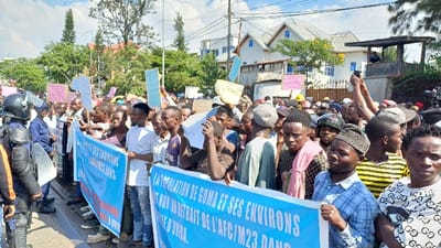 A daylight scene in Kabinzi, Rwanda, shows a dense gathering of individuals, predominantly adult males, participating in a protest on what appears to be a paved street. Two individuals dressed in black riot gear, including helmets, visors, body armor, and shields, stand on the far left, facing the crowd. The protestors hold numerous handheld signs of various colors, including pink, light blue, yellow, and white. A large light-blue banner with white text, partially visible, is held horizontally across the foreground by multiple individuals. The text on the banner includes phrases in French, such as "LA POPULATION DE GOMA ET SES ENVIRONS" and "NON AU RETRAIT DE L'AFC/M23 DANS." In the background, several multi-story buildings with white facades and diverse rooflines are visible, interspersed with trees and overhead utility lines. A person stands on a wooden observation structure attached to a wall on the right side of the image, while professional camera equipment on a tripod is also present among the protestors. Another individual in the crowd holds a white object, likely a phone or camera, aloft.