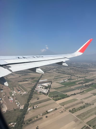 An aerial photograph captures a view from an airplane window, showcasing a segment of the aircraft's wing against a clear blue sky and the landscape below. The white wing, featuring a prominent red tip, extends from the upper left towards the bottom right of the frame. Visible on the underside of the wing are partial markings including "X A-VAO" and the Spanish phrase "NO CAMINE FUERA DE ESTA ÁREA" (Do not walk outside this area).
The background reveals an expansive rural and semi-urban area, identified as Santa María Zacatepec, Mexico. The ground is characterized by a mosaic of agricultural fields, varying in color from light brown to green, arranged in rectangular plots. These fields are interspersed with lines of trees, dirt roads, and scattered buildings, some of which appear residential while others suggest industrial or commercial structures with white rooftops. The scene is brightly illuminated, indicating daytime with clear weather conditions, and a few small, wispy clouds are visible in the blue sky. The overall perspective is tilted, suggesting the aircraft is in a bank or the photo was taken at an oblique angle.