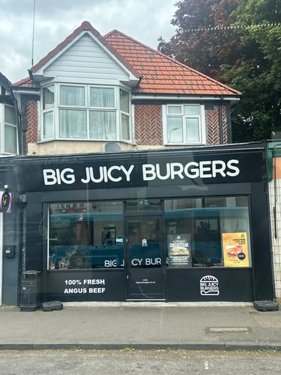 The image shows the storefront of "Big Juicy Burgers" in St Albans, United Kingdom. The store has a black facade with white lettering. The name "BIG JUICY BURGERS" is prominently displayed above the entrance. A menu board is visible in the window, advertising "LUNCHTIME JUST GOT JUICY!" for £8.95, and "PHILLY CHEESESTEAK" for £3.95. The restaurant's logo is displayed on the front. The store's address, "187", is visible on the building's exterior. A residential building is adjacent to the restaurant, with a red tiled roof and a bay window. A trash receptacle is adjacent to the main building.