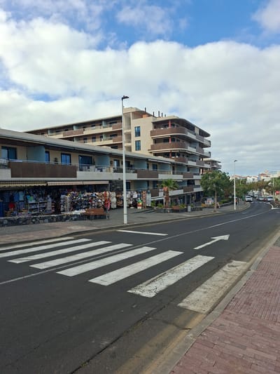 The image depicts a street scene in Santiago del Teide, Spain. In the foreground, a pedestrian crosswalk with white stripes is visible on an asphalt road. To the left of the crosswalk, a sidewalk paved with reddish bricks lines the street. Behind the sidewalk, a row of shops with awnings displays various goods, including clothing, hats, and bags. A wooden bench is placed in front of one of the shops.
Further back, a multi-story building with balconies and dark-framed windows dominates the scene. The building has a modern architectural style with curved balconies. Beyond this building, other structures and palm trees are visible, suggesting a coastal or resort area. The sky is partly cloudy with scattered white clouds against a blue backdrop. Cars are seen driving on the road in the distance. The overall impression is a commercial street on a day with fair weather.