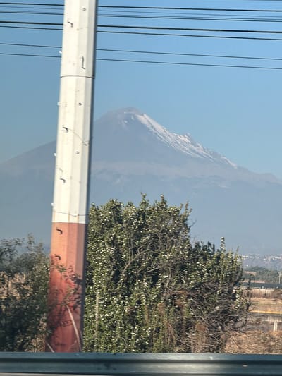 The image depicts an outdoor daytime scene in the vicinity of Santa María Zacatepec, Mexico, characterized by a clear sky and distant mountainous terrain.
In the background, a large, snow-capped stratovolcano is visible, partially obscured by atmospheric haze. Its upper slopes are covered in white snow, while the lower slopes appear grey-blue. The sky above and around the mountain is uniformly light blue. Faint, distant residential or urban structures are discernible at the base of the mountain on the far right.
In the midground, a multi-segment utility pole stands vertically, slightly left of the image's center. Its upper section is white/light grey, featuring several small, dark metal attachment hooks. The lower section of the pole transitions to a reddish-brown color. Multiple thin, dark electrical wires run horizontally across the upper portion of the sky, originating from or passing near the pole. To the right of the pole, a dense green bush with numerous small white blossoms occupies a significant portion of the mid-foreground. Sparse, drier brown vegetation is visible to the left of the pole and behind the flowering bush.
In the immediate foreground, a dark grey, metallic guardrail or barrier extends horizontally across the bottom of the frame. No individuals, animals, or specific actions are visible in the scene.