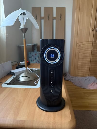The media file displays a close-up, rotated view of a modern black air purifier on a light wood surface, likely a desk or table. Behind the air purifier, a light wood cabinet with a grooved design is visible, suggesting a home or office setting. A crumpled light gray garment is draped over the cabinet.
The air purifier is the central object, featuring a circular LED display with blue illuminated text and segments indicating some form of measurement or setting, possibly related to air quality or fan speed. Below the display, a row of circular buttons with icons are visible, including one labeled "LIGHT" and another with a wave-like symbol.
In the foreground, to the left of the air purifier, a partially visible object with a translucent white plastic curved piece and a wooden handle is present. This object appears to be a disassembled fan or a component of a fan. It has a light bulb-like element at its center.
The overall lighting suggests daytime, with natural light illuminating the scene. The background is slightly out of focus, but the presence of the cabinet and the wooden furniture indicates an indoor environment. There are no people visible in the image. The location context is Idar-Oberstein, Germany.