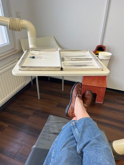 The image depicts the interior of a dental examination room in Monsheim, Germany, viewed from the perspective of a patient seated in a dental chair. The lower legs of the patient are visible in the foreground, crossed at the ankles, clad in blue denim jeans and brown leather loafers with gold-colored chain accents. The patient is positioned in a dark-colored dental chair, a portion of which is visible in the lower right.
In the mid-ground, a white medical cart on slender metal legs is situated next to the patient. It supports two rectangular stainless steel trays. The left tray contains a transparent syringe filled with a blue substance, accompanied by two white cotton rolls. The right tray holds a selection of sterile metallic dental instruments, including a dental mirror, various probes, and forceps. A white paper cup is mounted in a holder on the right side of the cart.
The background features white painted walls. A white window frame and a white radiator are visible on the left side of the room. An off-white, multi-segmented flexible arm, consistent with a dental light or X-ray unit, extends from the upper left. The floor is dark brown with a wood-patterned finish. A small, orange, two-step wooden stool is positioned beneath the medical cart. Behind the cart, partially obscured, is a decorative object resembling a stylized figure with a yellow body and dark, spiky hair. A white light switch panel is present on the far left wall. The scene is set for a dental procedure.