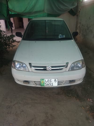 A white Suzuki Cultus car is parked in what appears to be an outdoor setting. The car is the primary object of focus, viewed from a slightly elevated angle, looking down on the front of the vehicle. The car is clean but shows some dust or dirt specks on its surface. The front grille, headlights, and the Suzuki logo are clearly visible.

Attached to the front of the car is a license plate with the text "LEC 15 1651" and "PUNJAB" written on it. The car is parked on a dirt or gravel surface, with some dry leaves and sparse vegetation visible around it. To the left of the car, a large section of green tarp or fabric is draped, creating a shaded area. In the background, part of a building with weathered walls is visible.

The overall lighting suggests it might be daytime, possibly late afternoon or early morning, given the shadow patterns and the ambient light. There are no people visible in the image, and the scene appears to be static, with the car simply parked. The location context provided is Haveli Jhangranwali, Pakistan. The image has a slightly tilted orientation.
