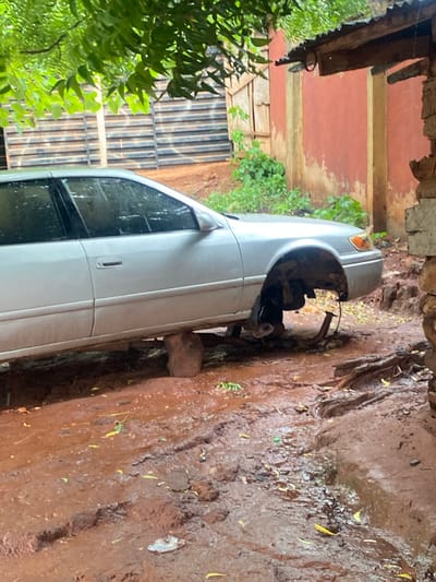 A light-colored sedan, possibly silver or white, is depicted stuck in a heavily muddy outdoor area. The image is rotated clockwise, showing the car resting on its side. The front passenger-side wheel of the car is missing, exposing the wheel well and suspension components. A large stone or block appears to be wedged under the car's frame near the front to support it or prevent it from sinking further into the mud.
The ground is a thick, reddish-brown mud, indicative of recent rainfall or consistently wet conditions, with small yellow leaves scattered across its surface and faint tire tracks. On the left side of the frame, vibrant green foliage partially obscures a building with reddish-brown walls where the paint is peeling. Wooden structural elements, including beams and a slatted fence-like structure, are visible around the building. An overhanging structure, possibly a roof or awning made of corrugated material, extends from the building.
The scene conveys a sense of a car breakdown or a difficult situation in a challenging, unpaved environment. No people are visible, and there is no visible text. The setting appears to be a less developed or rural area, consistent with the location context of Auchi, Nigeria.
