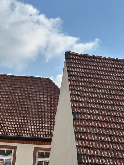 The image displays an architectural scene against a partially clouded sky in Offenbach an der Queich, Germany. The upper half of the frame is dominated by a clear, light blue sky with scattered white cumulus clouds. The lower half features two prominent tiled roofs. The roof on the right is a steeply pitched gable, covered in tightly arranged reddish-brown clay tiles, some exhibiting discoloration or dark staining. The vertical gable wall beneath this roof is painted a light beige or off-white. The very peak of this gable shows dark residue or material consistent with wear. To the left and slightly behind, a second, less steeply pitched roof is visible, also covered in similar reddish-brown clay tiles. Below this left roof, a section of a light beige building wall is present, featuring two partially visible windows with white frames and dark sills. No individuals or active subjects are present. The scene is static, depicting an exterior view of traditional European residential or commercial buildings under daylight conditions.