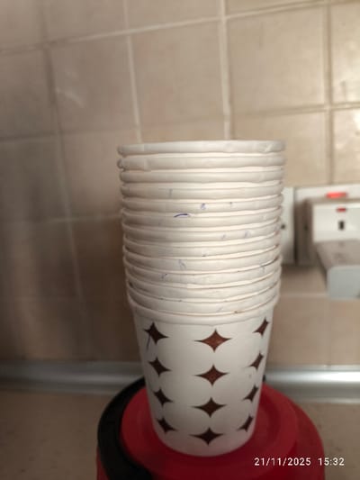 The image displays an indoor scene, located in North Nicosia, Northern Cyprus / Cyprus. The central focus is a vertical stack of approximately fifteen white disposable paper cups, each featuring a repeating pattern of dark brown, four-pointed star shapes on its lower section. Faint blue ink marks are visible on the upper rims and sides of several cups within the stack. The cups are resting on a red, circular object, which includes a black perimeter, situated on a light-colored countertop. A metallic trim runs along the junction between the countertop and the background wall. The wall is surfaced with light beige, square tiles, arranged in a grid pattern with light grout lines. To the right of the stacked cups, a white electrical wall outlet or a device plugged into it is partially visible; it exhibits an illuminated red indicator light and a connected white adapter. The environment is evenly lit. No individuals are present, and no actions or interactions are depicted. A digital timestamp in the lower right corner indicates "21/11/2025 15:32".