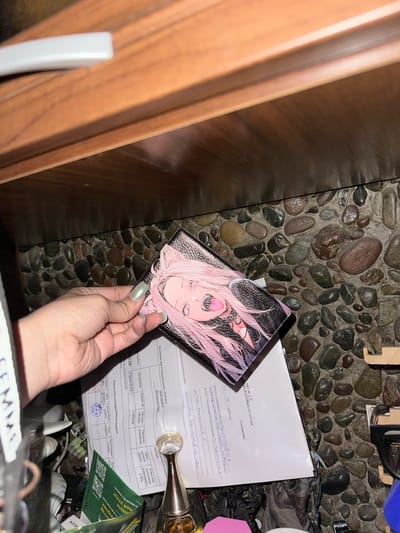 This media file captures a close-up, overhead shot of a hand holding a small, rectangular object decorated with an anime-style illustration of a pink-haired character. The hand, with its pale green, glossy fingernails, is positioned over a document with Russian text.
The document appears to be a form or report, with headings like "АКТ № 216" (ACT No. 216) and "ПАСПОРТ Водосчетчик" (PASSPORT Water Meter). Visible text includes dates such as "03 / 06 / 2026 год" (03 / 06 / 2026 year) and "18.05.26" (18.05.26), suggesting a focus on official documentation and potentially calibration or inspection of a water meter. Some handwritten entries are also present.
The background consists of a textured surface of smooth, rounded stones, typical of decorative landscaping or a natural setting. To the left, a portion of a wooden surface, possibly furniture or a wall, is visible. Other objects in the background are somewhat obscured but include what looks like a bottle with a metallic cap and a green package with Cyrillic text. The lighting suggests an indoor setting, possibly at night or in a dimly lit area, with a flash likely used for the photograph. The overall impression is one of a personal item and official paperwork being handled in a somewhat cluttered environment.