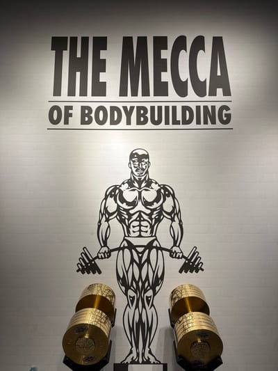 The image depicts an interior view of a fitness establishment in Serris, France. A light-colored, possibly white, wall with a brick-like pattern serves as the background. Centered on the upper portion of the wall, the phrase "THE MECCA OF BODYBUILDING" is displayed in large, black, sans-serif capital letters. Below this text, a prominent black graphic of a muscular male bodybuilder holding a barbell is affixed to the wall. The figure is stylized, emphasizing musculature. In the foreground, positioned on the floor and in front of the lower half of the bodybuilder graphic, are two large, golden, cylindrical objects. These objects resemble highly polished dumbbells or decorative weight stacks, each composed of multiple circular plates, mounted on dark, upright stands. No individuals are present in the frame.