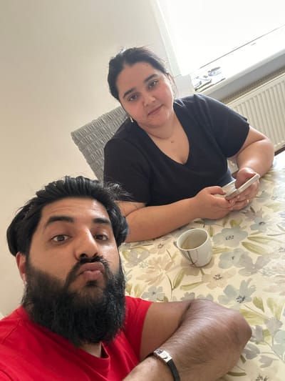 The image shows two people, a woman and a man, lying down. The woman is wearing a black t-shirt and is holding a smartphone, appearing to be engaged with it. She has dark hair and is looking towards the camera. To her right, a white mug with some liquid, possibly coffee or tea, is visible on a patterned surface that resembles a tablecloth or bedspread. The man, wearing a red t-shirt, is lying next to her. He has a beard and is also looking towards the camera. His arm, adorned with a black bracelet with a silver buckle, is visible. The background suggests an indoor setting, possibly a bedroom or living room, with a radiator visible in the upper left corner. The lighting indicates it is daytime. The overall impression is a casual, relaxed moment shared between the two individuals, possibly at home.