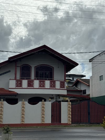 The image displays a multi-story building, likely a house, under an overcast sky. The building features a distinctive architectural style with a prominent, steeply sloped, dark red-brown tiled roof and white-painted walls. Decorative elements include red-brown intricate lattice patterns on balcony railings and carved details around arched windows. Two notable arched windows are visible on the upper floor, with further decorative panels flanking them.
Utility lines crisscross the grey, cloudy sky above the building. To the right of the main structure, a paved road or driveway is visible, bordered by a fence. This fence incorporates dark red-brown horizontal panels and sections with light-colored, patterned stone or tile work. Glimpses of other buildings, also with white facades, are present in the background, suggesting a built-up area.
The scene is static, with no people or significant activity taking place. The time of day is daytime, characterized by the bright, diffused light, but the sky is entirely covered with grey clouds, indicating overcast weather. Small patches of green vegetation are visible near the road. While there appear to be small rectangular signs or plaques on the building walls, any text on them is unreadable due to blurriness. The setting is Matara, Sri Lanka.