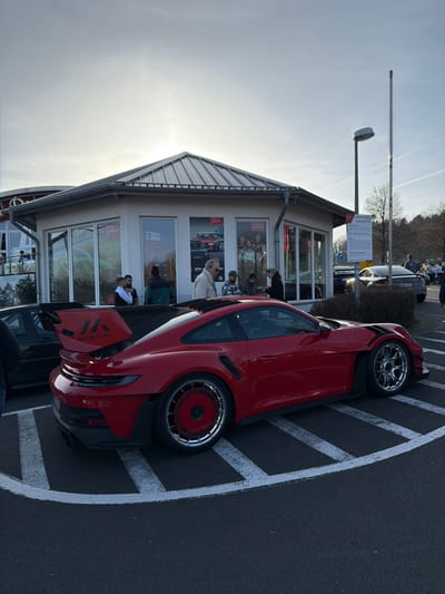 The scene depicts an outdoor gathering at a commercial building in Meuspath, Germany, under a bright, partly cloudy sky. In the immediate foreground, a modified red Porsche 911 GT3 RS (992 generation) is parked, angled towards the left. The car features a large black rear spoiler with white "MK MANTHEY" branding. Its rear wheels are multi-spoke with a prominent red, circular, disc-like cover, while the front wheels are polished silver multi-spoke. The vehicle rests on dark asphalt, partially outlined by white striped parking bay markings forming a semi-circular pattern.
Behind the red Porsche, a dark-colored car with a large rear wing is partially visible to the left. The background features a single-story building with a light-colored facade and a dark grey, corrugated metal hip roof. The building's large windows display automotive-related advertisements, including a green sports car, classic vehicles, and lifestyle imagery. Approximately 10-15 individuals, casually dressed, are gathered in front of and near the building's entrance, engaged in conversation or observation. One man in a light-colored jacket is prominently visible among the group.
To the right of the building, a light pole with a street lamp stands, accompanied by a white informational sign on a pole, below which are blue circular road signs. In the far background, additional parked vehicles, including a dark Audi TT, are visible alongside bare trees.