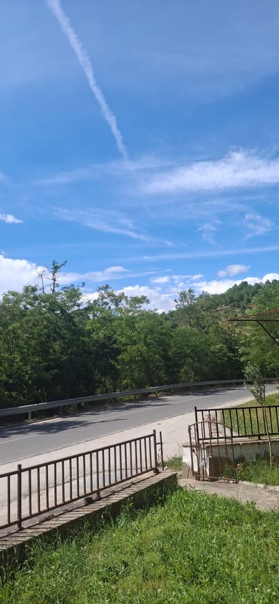 This is a vertical, eye-level, outdoor photograph of a road winding through a green, hilly landscape under a bright blue sky with wispy white clouds. In the foreground, there is a metal fence with vertical bars and some decorative scrollwork on the right side. Beyond the fence is a concrete walkway and a patch of green grass. A paved road runs parallel to the fence, separated by a silver metal guardrail. The road curves to the left and disappears into a dense forest of green trees. The trees cover the slopes of the hills, which rise in the background. A thin contrail stretches across the sky from the upper left to the lower right. The scene appears to be a rural or semi-rural area.