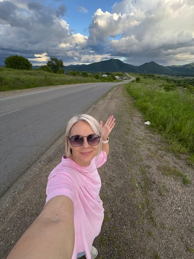 A woman with blonde hair and sunglasses takes a selfie on the side of a rural road. She is wearing a pink t-shirt. The road curves ahead, disappearing into a valley with rolling hills and mountains in the distance. The sky is partly cloudy with dramatic cloud formations. The area appears to be a natural, undeveloped landscape. There is no indication of the city of Теплоозёрск, Russia, in the image.