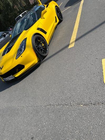 The image shows a bright yellow sports car parked on an asphalt surface. The car is oriented with its wheels pointing towards the left side of the frame, and the image is rotated 90 degrees counter-clockwise. Yellow lines are painted on the asphalt, suggesting a parking lot or a road with designated markings. The car features black rims and a black trim around the headlights and front grille. Part of a license plate is visible on the front of the car, displaying the text "IB J794". The setting appears to be outdoors during the daytime, as indicated by the bright sunlight and the shadows cast on the asphalt. The weather seems clear and sunny. In the background, there are glimpses of green foliage and possibly other parked vehicles. The overall scene conveys a sense of stillness, with the focus primarily on the car.