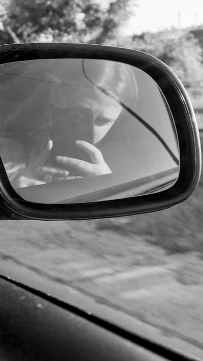 A black and white, close-up shot shows the reflection of a person taking a selfie in a car's side-view mirror. The person's face and hands are visible in the reflection, holding a dark-colored smartphone. Outside the mirror, blurry trees and a hillside are visible, suggesting the car is in motion on a rural or semi-rural road, potentially in or near Străisteni, Moldova. The foreground shows the car's interior bodywork and the edge of the side-view mirror housing. The overall impression is a candid, in-the-moment capture.
