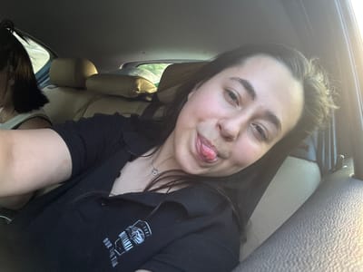 A young female is depicted in a selfie taken inside a vehicle, located in Omarchevo, Bulgaria. She is positioned in a passenger seat, with her head tilted to the side and her tongue playfully extended from her mouth. She has dark hair and wears a black, short-sleeved collared shirt that features a white logo and text on the left chest, partially reading "BELMANIA 2024 OUR THE TIM." A silver chain necklace with a small heart-shaped pendant is visible around her neck. The car interior shows beige-colored upholstery on the seats and headrests. In the background to the left, the back of another person's head with dark hair and a white top is partially visible, indicating a second occupant in the vehicle. Strong natural light illuminates the right side of the frame, suggesting daytime, and green foliage is faintly visible through the vehicle's rear window.