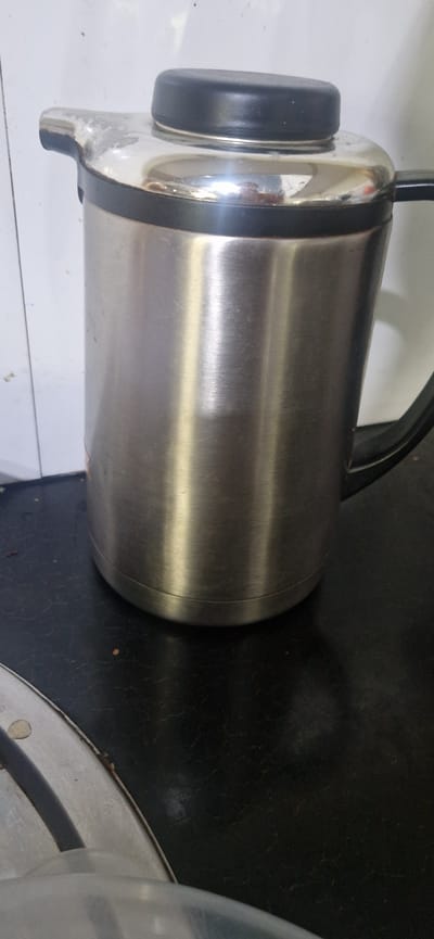 A stainless steel thermos with a black lid and handle sits on a dark, textured surface. The thermos is cylindrical and appears to be empty. The background is a white tiled wall. The image was taken in the city of Suzak, Kyrgyzstan.