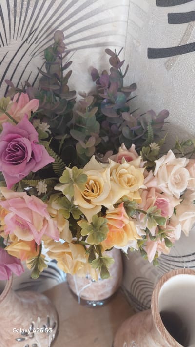 This is a medium shot, top-down photograph of a bouquet of artificial flowers. The flowers are a mix of roses and greenery, in shades of pink, yellow, and purple. The bouquet is arranged in a vase that is partially visible in the upper right corner of the frame, with another similar vase in the lower right. The background is a wall with a textured, off-white surface and a pattern of black and grey lines.
The lighting suggests it is daytime, but the overall mood is neutral, with no strong emotional cues. The image seems to be taken indoors, possibly in a home or decorative setting. The focus is primarily on the flowers, with the vases and background being secondary.
There is a visible text watermark in the bottom right corner of the image: "Galaxy A36 5G".