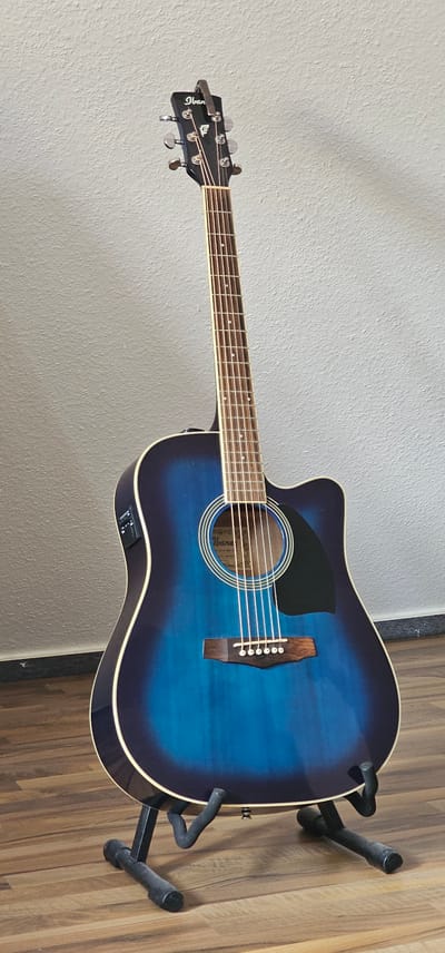 An acoustic-electric guitar with a blue sunburst finish stands upright on a black guitar stand. The guitar has a cutaway body and is positioned in front of a textured, light-colored wall. The floor is made of wood planks. The scene is indoors, and there are no people present. The guitar is the sole subject of the image. The overall impression is one of a static display in what appears to be a room or studio setting. The image does not contain any information that would indicate its location in Albertshausen, Germany.