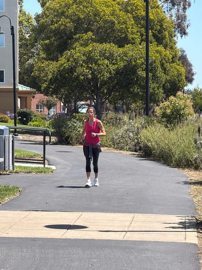 This media file is a vertical, full-body shot of a woman exercising on a paved path in what appears to be a park or residential area. The woman is wearing a bright pink tank top, black leggings, and white athletic shoes. She is positioned mid-stride, suggesting she is running or jogging. Her body is angled towards the viewer, and she is looking slightly off-camera.
The scene is outdoors, with ample greenery and trees surrounding the paved path. The path itself is a dark asphalt with a lighter-colored concrete divider running alongside it. Shadows are cast on the path, indicating it is daytime, likely mid-morning or afternoon due to the brightness and angle of the sun. The weather appears to be clear and sunny, with a bright blue sky visible in the background.
In the background, there are glimpses of buildings, one appearing to be made of red brick with white-trimmed windows, and another lighter-colored building with windows. There is also a black metal bench or rack on the side of the path. The overall atmosphere suggests a healthy and active lifestyle, with the woman enjoying an outdoor workout. There is no visible text in the image.