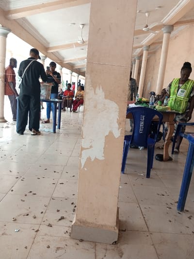 The image depicts a public gathering area in Ajalli, Nigeria, characterized by a light-colored tiled floor and a colonnaded architectural structure with a series of pillars and arches supporting a ceiling equipped with multiple white ceiling fans. A prominent, light-beige square column with extensive peeling paint dominates the right foreground, positioned centrally within the frame. The tiled floor is visibly soiled with numerous small, dark, organic debris, possibly peanut shells, and scattered pieces of trash.
In the midground, to the left of the central column, a person in a black long-sleeved shirt and dark trousers stands facing away from the camera, positioned at a light blue plastic table. On this table rests a white container, consistent with a ballot box or a registration bin. Opposite this individual, a person in a dark t-shirt and blue jeans holds a black smartphone, aiming it towards the white container, indicating photographic or video recording. Another person in a brown collared shirt and dark trousers stands further to the left. Behind this immediate interaction, several individuals, predominantly women in colorful attire, are seated on red plastic chairs, facing towards the activity.
To the right of the central column, an individual identified by a neon green reflective vest with visible "INEC" branding (Independent National Electoral Commission) over a patterned skirt stands beside a light brown wooden table. The individual's head is bowed. On the table are several plastic water bottles, green containers, and assorted papers. A blue plastic chair is positioned in front of this table. Other individuals are partially visible seated behind the INEC official's table.