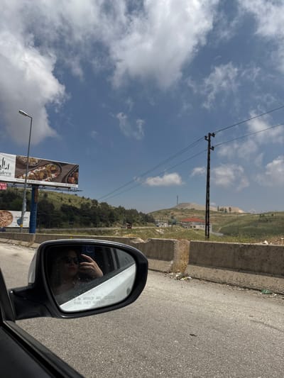 A car's side mirror reflects a person wearing sunglasses and a ring on their finger, holding up a smartphone. The mirror displays text that reads "OBJECTS IN MIRROR ARE CLOSER THAN THEY APPEAR." Outside the car, a billboard advertises a restaurant named "AL ALEEN RESTAURANT" with images of food. The scene is set in Dahr El Baidar, Lebanon, under a bright, cloudy sky. The landscape features rolling green hills dotted with trees and some buildings. Power lines and a utility pole are visible in the background, along with a concrete barrier along the side of the road. The road itself appears to be asphalt with some debris scattered along the roadside.