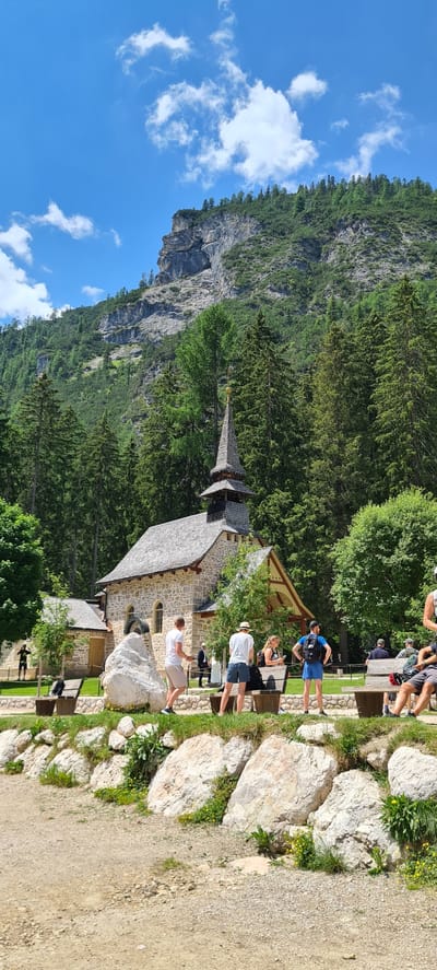 The image depicts a scenic view of a stone church nestled amidst lush green trees and a towering, rocky mountain. Several people are visible, engaged in various activities. In the foreground, a person in a white t-shirt and shorts is leaning against a large rock, seemingly taking a break. Further back, a group of people are gathered, some standing and others seated at wooden benches. One individual is wearing a blue backpack, suggesting they might be hikers. The scene appears to be set during daytime, with bright sunlight casting shadows. The weather seems pleasant, indicated by the clear blue sky with scattered white clouds. The presence of the church and the natural landscape suggests a tourist or recreational area, likely a hiking trail or a scenic viewpoint in the Prags - Braies region of Italy. There is no visible text in the image.