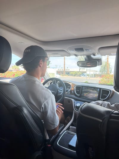 The image shows a man sitting in the driver's seat of a car. He is wearing a grey t-shirt, a baseball cap, and sunglasses. He is holding the steering wheel, and a purple object is visible in his hands. The car is parked on a street in Palm Desert, California. The exterior view through the windshield shows a street with palm trees and buildings. A storefront sign with the words "RUCCI & ROOM" is partially visible. A vehicle backup camera display is visible in the dashboard.