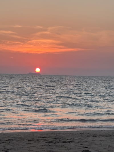 This image depicts a sunset over the ocean in Phra Ae, Thailand. The sky is dominated by hues of orange and red, transitioning from a pale peach at the top to vibrant orange and deeper red tones near the horizon. Sparse, elongated clouds are visible in the upper and middle sections of the sky. The sun, a distinct, circular, bright orange-red orb, is positioned centrally on the horizon, appearing to rest directly above a dark, low-lying island or landmass. The sea, occupying the middle ground, exhibits a dark blue-gray color with moderate ripples and reflections of the sunset's orange-pink light. In the foreground, a sandy beach, appearing dark gray, stretches across the bottom of the frame, with gentle waves breaking at its edge, creating a narrow strip of orange-red reflection where the water meets the sand. No individuals or man-made objects are present in the scene.