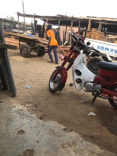 A red and white motorcycle stands on a dirt and concrete surface near a building with lumber. A person wearing an orange shirt and blue pants pushes a wooden cart laden with lumber. A truck with Chinese characters is visible in the background. Tubes or pipes are in the lower left corner. The scene appears to be outdoors, potentially a marketplace or industrial area.