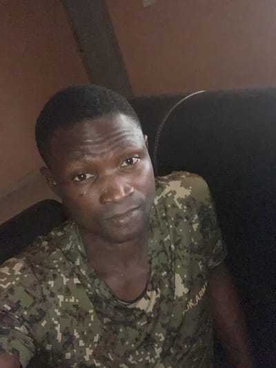 An adult male subject is depicted in a close-up, self-portrait style photograph, situated in an unspecified indoor location within Kado, Nigeria. The subject, who has dark skin and short dark hair, is wearing a short-sleeved digital camouflage t-shirt with the text "JK ARMY" visible on the right chest area. A faint mark or scar is present on his left cheek. He is looking directly at the camera with a neutral expression. The background is dimly lit and indistinct, showing a dark, possibly upholstered, surface to the subject's right and light brown or tan walls.