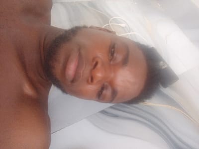 A selfie captures a shirtless man with dark skin and a beard, lying on a tiled surface. He gazes into the camera with a neutral expression. White earphones are visible near his head. The background is a blurred pattern of white and gray tiles, creating a stark contrast with the man's skin tone. The lighting is soft, emphasizing the contours of his face and upper body.