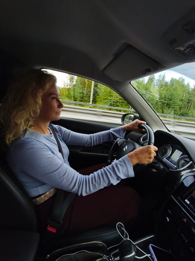 An outdoor, eye-level, medium shot shows a woman driving a vehicle on a highway. The woman, who has blond, curly hair, is wearing a light blue long-sleeved shirt and a reddish-brown skirt. She is holding the steering wheel with both hands, looking forward. The interior of the car is visible, including the dashboard with gauges and a screen, and the black leather seats. Outside the car, a forest of green trees lines the highway. The weather appears to be sunny. The image was taken in Kissolovo, Russia.