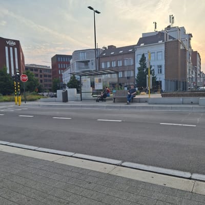 The image captures a street scene in Mechelen, Belgium, likely during late afternoon or early evening given the lighting. In the foreground, a paved road with white lane markings is visible, bordered by a textured gray sidewalk and a granite-like curb. Beyond the sidewalk, a bus stop shelter with a glass roof and metal supports stands.
Two individuals are seated on a bench at the bus stop. One, a person wearing a dark shirt and blue jeans, is looking to the right. To their left, another person, wearing a red shirt and dark pants, is seated and appears to be attending to a stroller or infant carrier in front of them, which contains a baby. The bus stop also features a trash receptacle.
In the background, a multi-story building with a modern facade, featuring a prominent "VDAB" logo, is visible on the left. Other buildings with varied architectural styles, including brick facades and white exteriors with numerous windows, line the street. Several communication antennas are mounted on the rooftop of one of the buildings. A street lamp with two lights stands tall near the bus stop. A red stop sign with white lettering is prominently displayed on a traffic signal pole to the left of the bus stop. The sky above is partly cloudy, with a warm, diffused light suggesting sunset or sunrise.