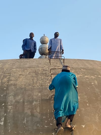 Three individuals are depicted on and around a large, weathered concrete or stucco dome structure in the city of Barako, Nigeria, under a clear blue sky.
In the foreground, an individual viewed from behind is actively ascending a fixed metal ladder attached to the dome's exterior. This person wears a teal-blue long tunic over black trousers, tan sandals, and a striped knitted cap.
Atop the dome, two other individuals are standing. To the left, a younger individual in a blue button-up shirt and dark trousers looks towards the camera. Partially obscured behind this individual is another person's head and arm. To the right, an older individual wearing a light blue traditional long garment and bright green sunglasses stands, looking slightly upward and to the right.
Between the two standing individuals on the dome's surface, a metallic finial ornament consisting of two stacked spherical elements topped with a spike is visible. The dome itself exhibits surface cracks and discoloration. The scene is illuminated by natural daylight.