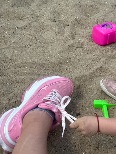 In Donchovci, Bulgaria, on a sandy surface that appears to be a playground or beach, a close-up shot captures a child's hand and a portion of an adult's leg and shoe. The adult's leg is clad in a grey sock and a bright pink athletic shoe with white laces. The child's small hand, covered in sand, is actively engaged in holding and manipulating the white shoelace of the adult's shoe. To the right of the adult's shoe, a metallic pink toy, possibly a mold for sandcastles, sits on the sand. Further to the right and slightly lower, a bright green plastic toy hammer rests on the sandy ground. A thin red string bracelet with a small, golden charm is visible on the child's wrist. The overall environment is characterized by the grainy texture of sand and hints of natural light creating subtle shadows.