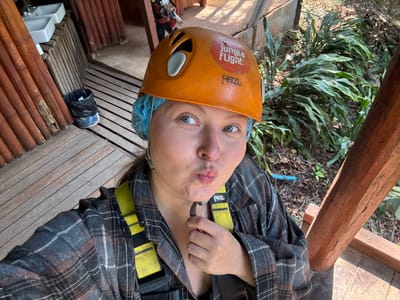 An eye-level portrait shows a person in an outdoor setting, positioned as if hanging upside down, though the image itself is rotated. The person, who appears to be an adult with fair skin and light-colored eyes, is looking directly at the camera with a playful expression, her lips pursed in a 'fish face'. She is wearing an orange safety helmet with white accents, underneath which a light blue, disposable hair net or cap is visible. A sticker on the helmet reads "JUNGLE FLIGHT" with a graphic of a bird. The brand "PETZL" is also visible on the helmet.
The person is wearing a grey and brown plaid long-sleeved shirt and a yellow safety harness, also bearing the "PETZL" brand name. The harness straps are visible across her shoulders and chest.
The background suggests an adventure activity in a jungle or forest environment. To the left (from the viewer's perspective, assuming the image is correctly oriented), there's lush green foliage with large leaves, indicative of tropical vegetation. A thick wooden beam, part of a structural element like a platform or tower, is prominent on this side. To the right, there's a wooden platform or structure, possibly part of a zipline station or observation deck, made of horizontal wooden planks. Further right and below, there are vertical wooden poles or slats, and some white sink basins are partially visible, suggesting a facility area. A black trash bag and a blue container are also seen on the wooden platform. The lighting suggests it is daytime.