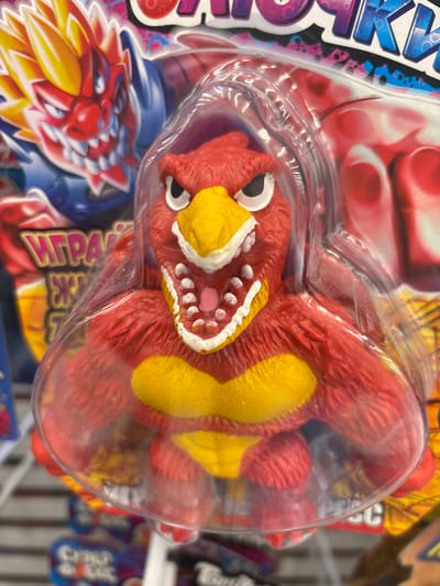 The image shows a close-up, eye-level shot of a red, fuzzy toy monster with a yellow beak and chest. The toy is packaged in clear plastic, which is slightly creased. The monster has large white eyes with black pupils, sharp white teeth, and a pink tongue. Its mouth is open, revealing its teeth and tongue. Behind the toy, the packaging displays graphics of a red and yellow dragon-like creature with fiery hair and blue armor, and text that appears to be in Cyrillic. The background is blurred, suggesting a retail environment with shelves and other products. The overall lighting is bright and even. No specific location in Kuzmolovsky, Russia, is visible in the image.
