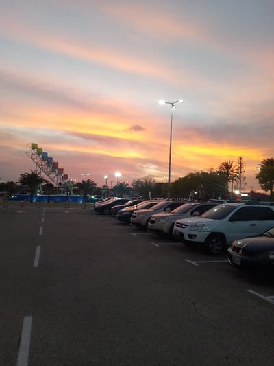 This image depicts an outdoor asphalt parking lot at dusk in La Cruz del Pastel, Venezuela, under a sky displaying vivid orange, yellow, and pink sunset hues. Multiple streetlights and facility lights are illuminated.

The foreground features dark asphalt with white dashed lines marking parking spaces. A row of approximately 15-20 parked passenger vehicles, predominantly SUVs and sedans in colors such as white, silver, black, and dark blue, occupies parking bays on the right side of the frame.

A tall, illuminated streetlight with a dual-lamp fixture stands in the mid-ground. Palm trees and other dense green foliage are visible across the mid-ground and background. To the left, a large metal structure consistent with a water park attraction is present, featuring multiple brightly colored (blue, green, yellow, orange, red) ascending segments resembling water slides or collection buckets. Blue accent lighting is discernible near its base. Several communication towers or utility structures are visible on the far right horizon. No individuals are present in the scene.