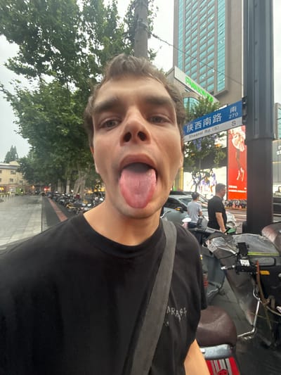 A man is featured in a close-up selfie, making a playful expression with his tongue sticking out. He has light skin and brown hair, and is wearing a black t-shirt with the partial text "ARAS" visible on it. The photo is taken on a city street during what appears to be an overcast day.
In the background, a blue street sign clearly identifies the location as "陕西南路" (Shaanxi Nan Lu), also labeled "Shaanxi Rd. (S) 270 S", confirming it as Shaanxi South Road in Xuhui District, China. A modern building with reflective glass windows stands prominently behind the street sign, and a green sign with "PARISON" (likely part of "PARKSON") is also visible.
The street is lined with green trees. Other people can be seen in the middle ground, alongside parked motorcycles or scooters and a partial view of a car. A large red billboard is also visible further down the street. The overall setting is a bustling urban environment.