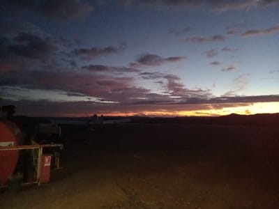 The image captures an airfield or private airstrip in Santa Juana, Chile, during a twilight period, evidenced by the low light and sky coloration. In the foreground-left, a large, cylindrical, red storage tank with visible piping and a pressure gauge is situated. Adjacent to this tank, a red cabinet or box, identified by a white fire extinguisher pictogram, is mounted on a metal frame. In the midground, on a dark, unpaved surface, the forms of at least two light aircraft or gliders are discernible as dark silhouettes against the lighter horizon. The background features a low, undulating horizon line, suggesting hills or dense foliage. The sky above transitions from deep blue and purple overhead to a prominent band of orange and yellow light directly above the horizon. Scattered dark clouds are present throughout the sky. No individuals or active movements are visible.